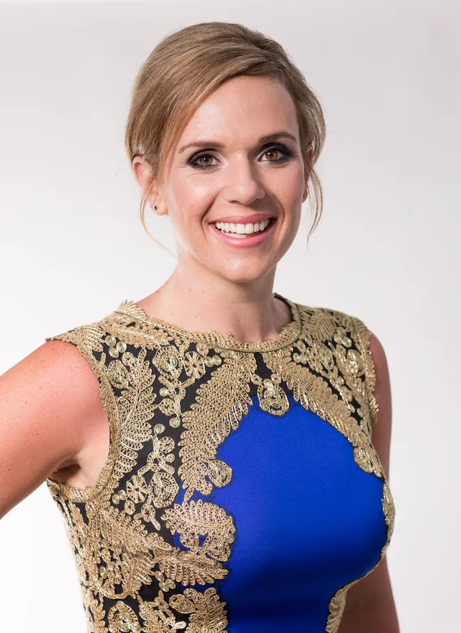 Woman in blue dress with intricate gold lace detail, smiling at the camera against a neutral background.