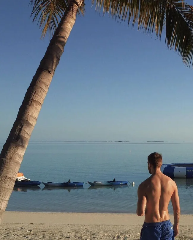 Man in blue shorts on a sandy beach, gazing at calm ocean with moored boats under clear sky.