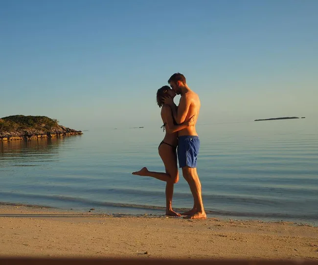 A couple embraces and kisses on a serene beach at sunset with clear skies and calm water.