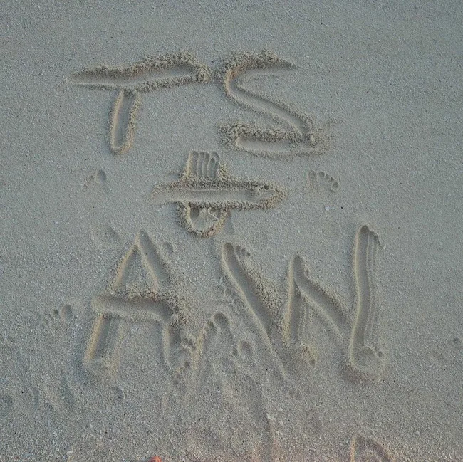"Letters 'TS + AW' written in sand with visible footprints around them on a beach."