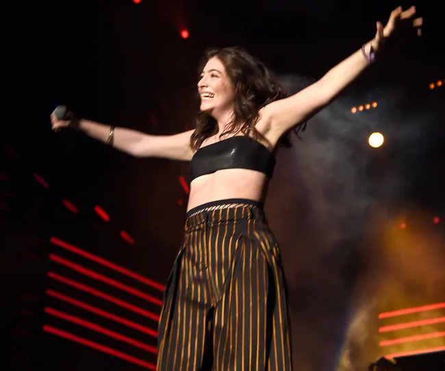 A performer on stage with arms outstretched, wearing a black top and striped pants, holding a microphone.