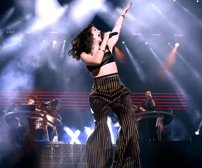Lorde performing energetically on stage with vibrant lights and musicians in the background.