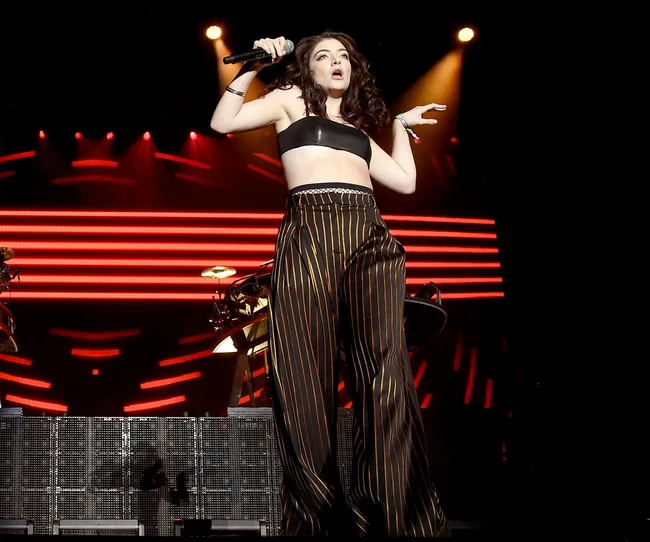 Singer performing on stage with striped pants and a black crop top, holding a microphone, red lights in the background.