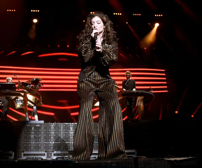 Singer performing on stage in striped outfit, dramatic stage lighting, musicians and instruments in the background.
