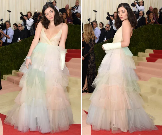 Lorde at Met Gala in pastel tiered gown with cast on right arm, posing on red carpet with photographers in background.