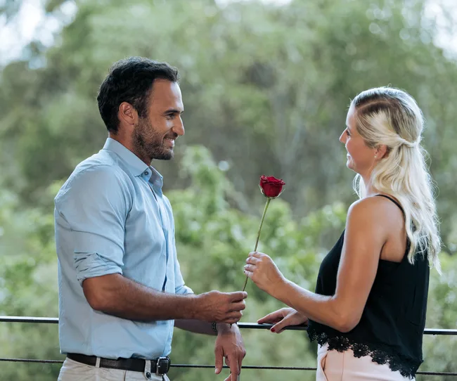 Man in blue shirt giving a red rose to a woman in black top, both smiling in an outdoor setting.