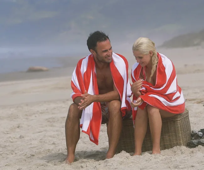 Couple sitting on the beach wrapped in red and white striped towels, smiling and talking.
