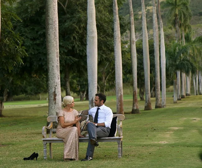 A dressed-up couple sits on a park bench, surrounded by tall trees, engaged in conversation with shoes off to the side.