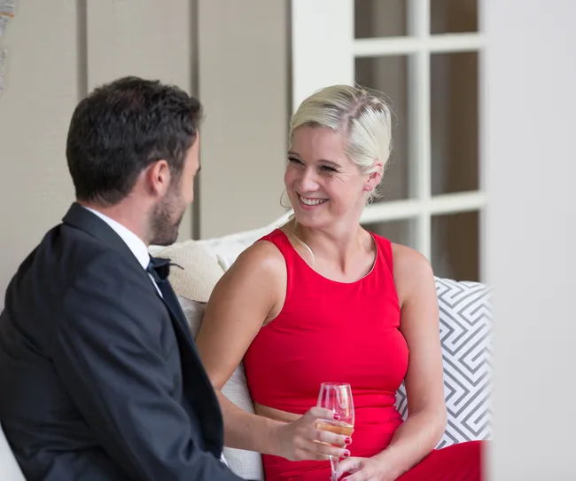 A woman in a red dress smiles while holding a glass of champagne, sitting with a man in a tuxedo.