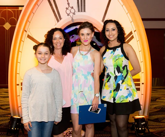 A group of four people smiling in front of a giant clock backdrop, with three women and a girl in casual and colorful attire.
