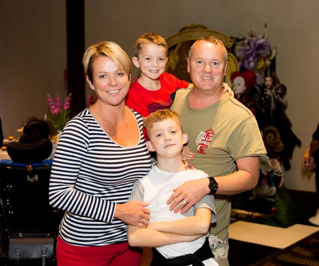 A family of four posing indoors, two adults and two children smiling, with a decorative background.
