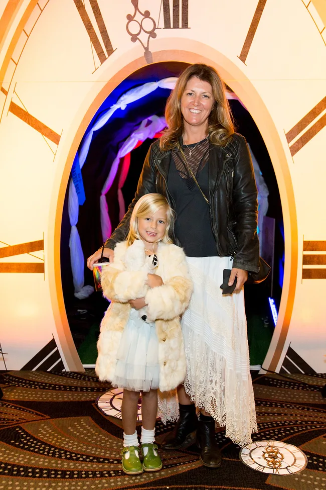 Woman and child in white outfits, standing in front of a large clock backdrop. The child holds a drink and a plush toy.