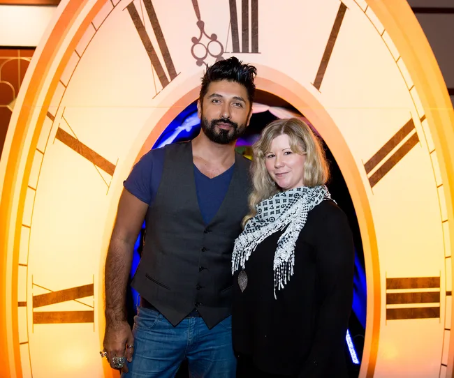 Two people stand in front of an oversized clock backdrop, smiling at the camera.
