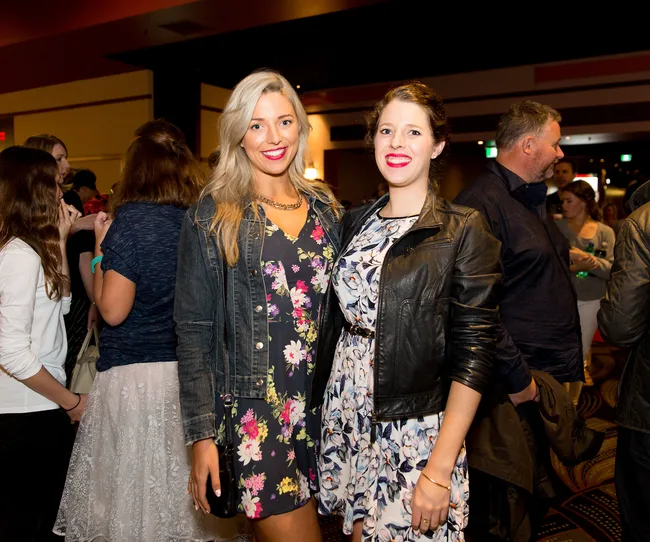 Two women in floral dresses and jackets smiling, standing in a crowded indoor gathering.