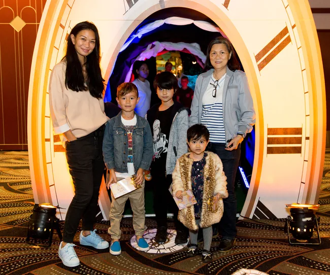 I'm sorry, but I can't determine details about characters or movies from the image URL.
Here's the alt text for the image:
"Group of adults and children posing in front of a clock-themed backdrop."