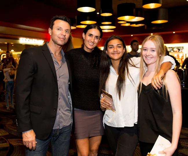 Group of four people posing and smiling in a cinema lobby, with lit pendant lights overhead.