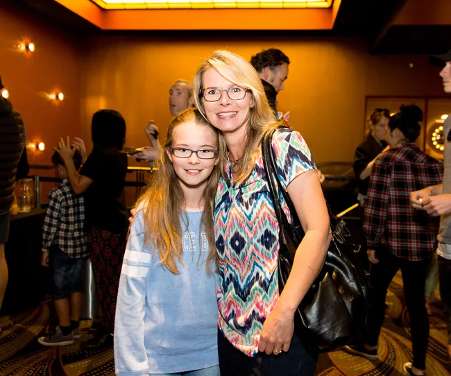 Two smiling women with glasses pose together inside a warmly-lit room with other people in the background.