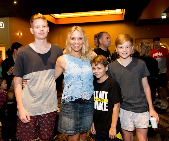 A woman is posing with three boys in a crowded indoor setting, smiling at the camera.