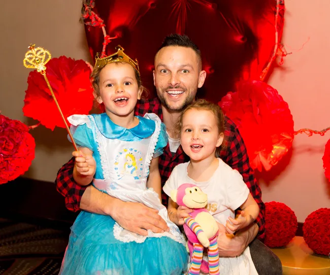 Father and two children, one in an Alice costume with a crown and wand, smile in front of vibrant red decor.