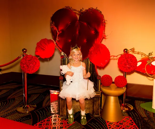 Child in a white dress and gold crown sits on a throne with a heart-shaped backdrop, holding a teddy bear.