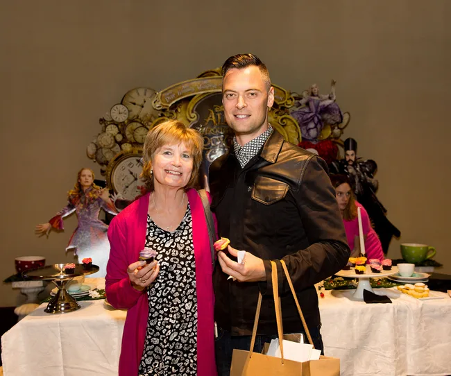 Two people stand smiling at a tea party event with "Alice Through the Looking Glass" decorations in the background.