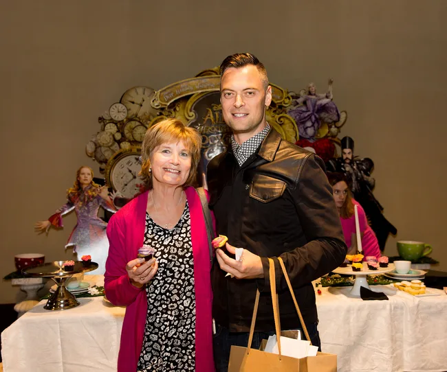 Two people stand smiling at a tea party event with "Alice Through the Looking Glass" decorations in the background.