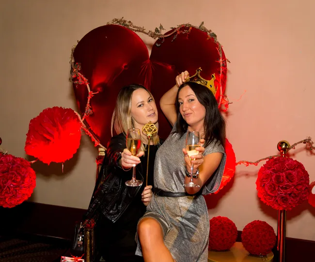 Two people celebrating, holding glasses of champagne, in front of a heart-shaped backdrop with red decorations.