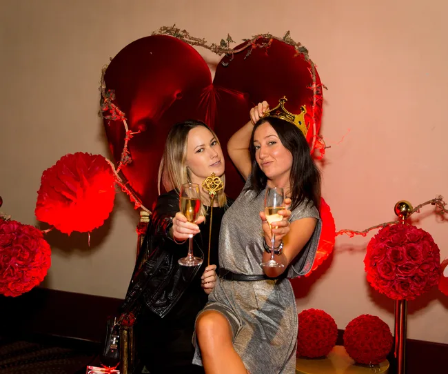 Two people celebrating, holding glasses of champagne, in front of a heart-shaped backdrop with red decorations.