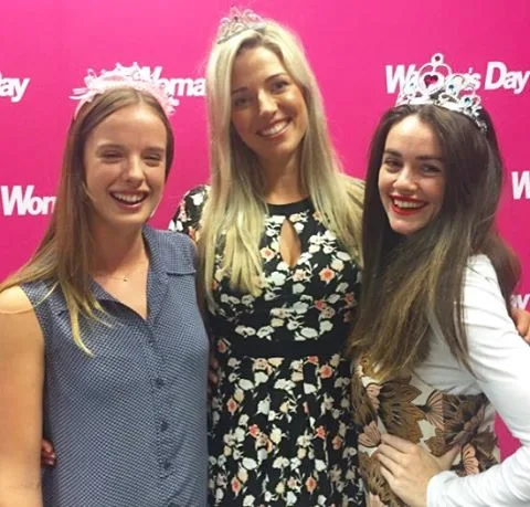 Three women in tiaras smiling in front of a pink "Woman's Day" backdrop.