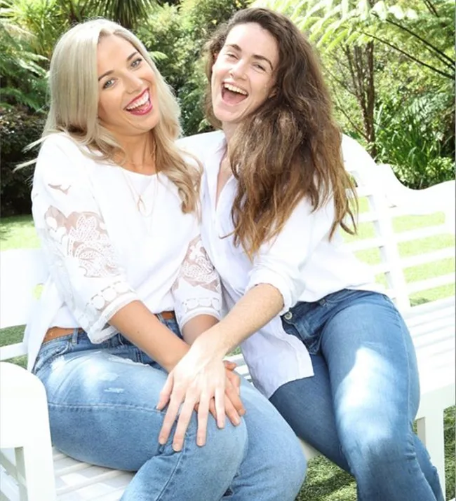 Two women smiling and sitting on a white bench outdoors, both wearing white tops and jeans, surrounded by greenery.