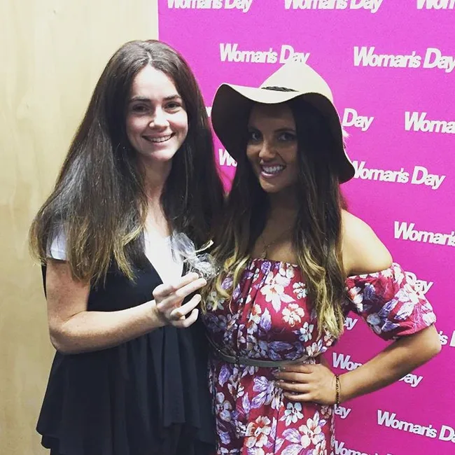Two women smiling in front of a "Woman's Day" backdrop; one holding a small item, the other wearing a floral dress and hat.