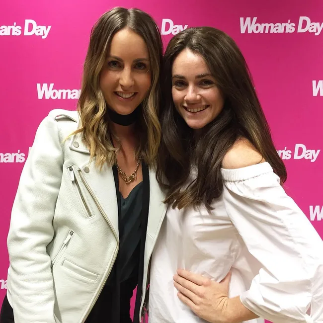 Two women smiling in front of a pink "Woman's Day" backdrop, one in a white jacket, the other in a white top.