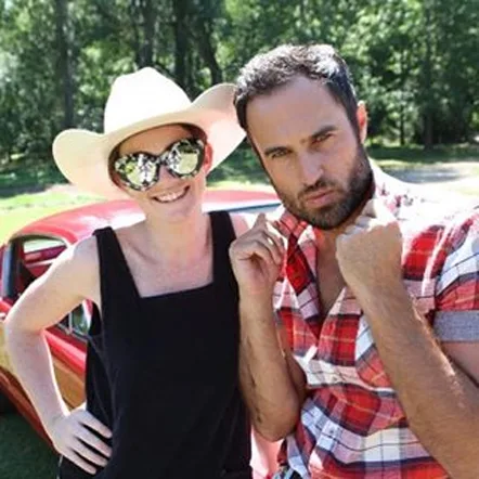 Two people posing outdoors; one in a cowboy hat and sunglasses, the other in a plaid shirt, with a classic car behind them.