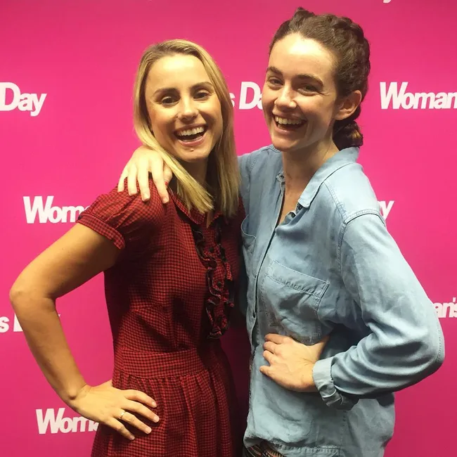 Two women smiling and posing together in front of a pink "Woman's Day" backdrop.