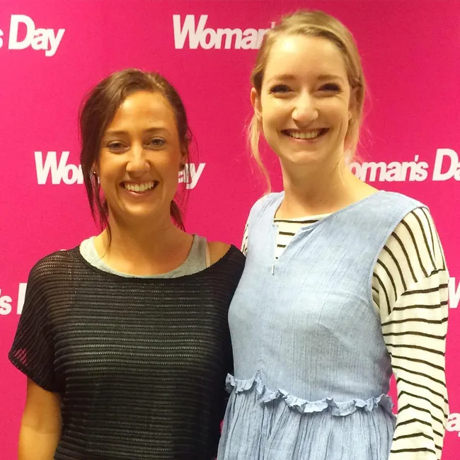 Two smiling women in front of a pink "Woman's Day" backdrop.