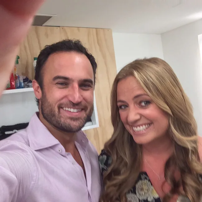 Two people smiling and taking a selfie indoors; the man wears a pink shirt and the woman has long, wavy hair.