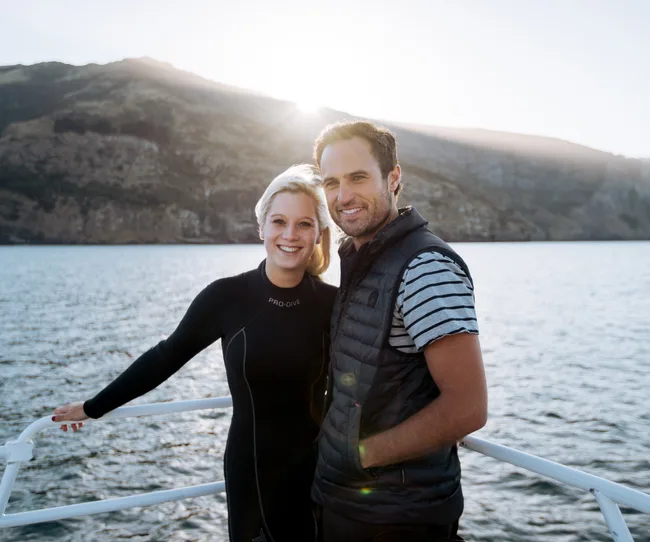 A man and woman smiling on a boat, with mountains and water in the background at sunset.