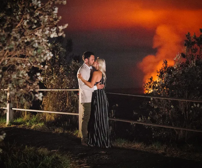 A couple embraces near a railing with a red-orange sky and smoke in the background, surrounded by foliage.