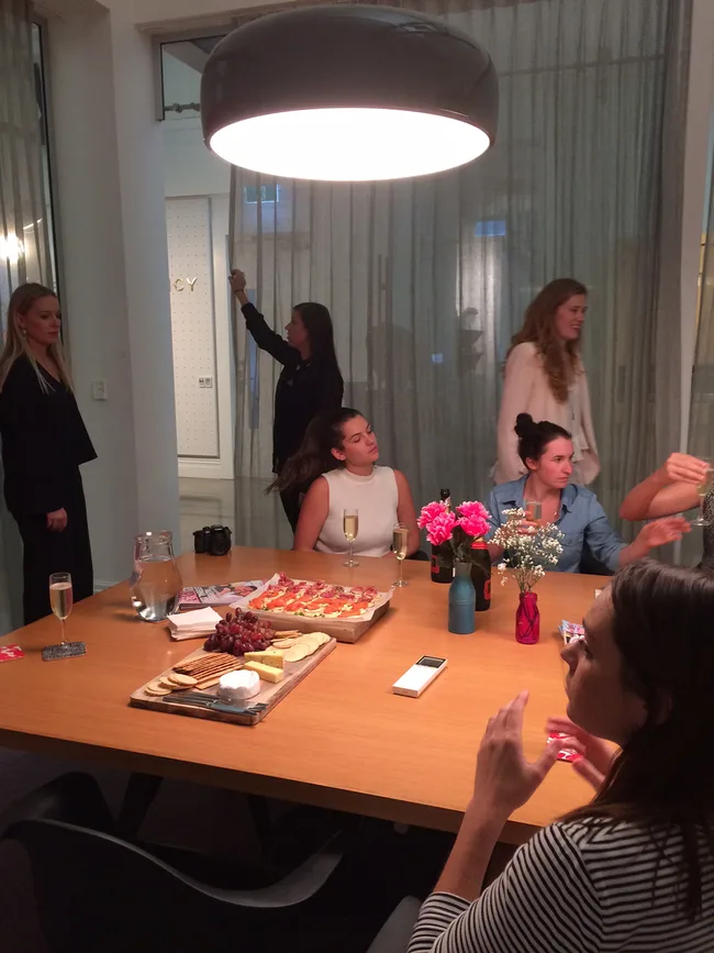 A group of people sitting around a table with appetizers, flowers, and drinks under a large hanging light.