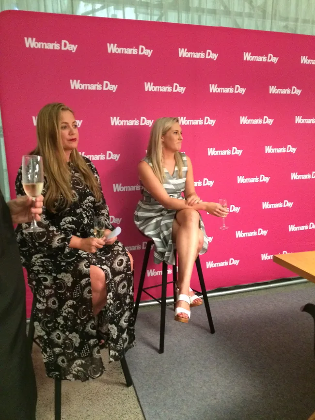 Two women sitting on stools, holding drinks, in front of a pink "Woman's Day" backdrop.