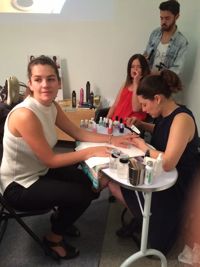 Manicure session in progress; woman receiving nail care while another gets her hair styled by a male stylist in a salon.