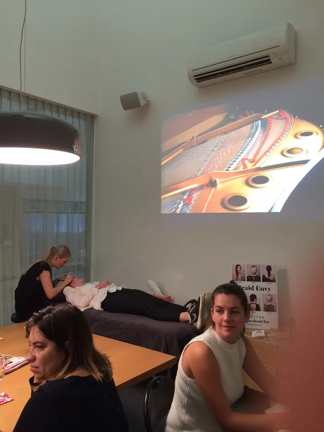 Group of women in a room; one lies on a table, another attends her. A piano image is projected on the wall.