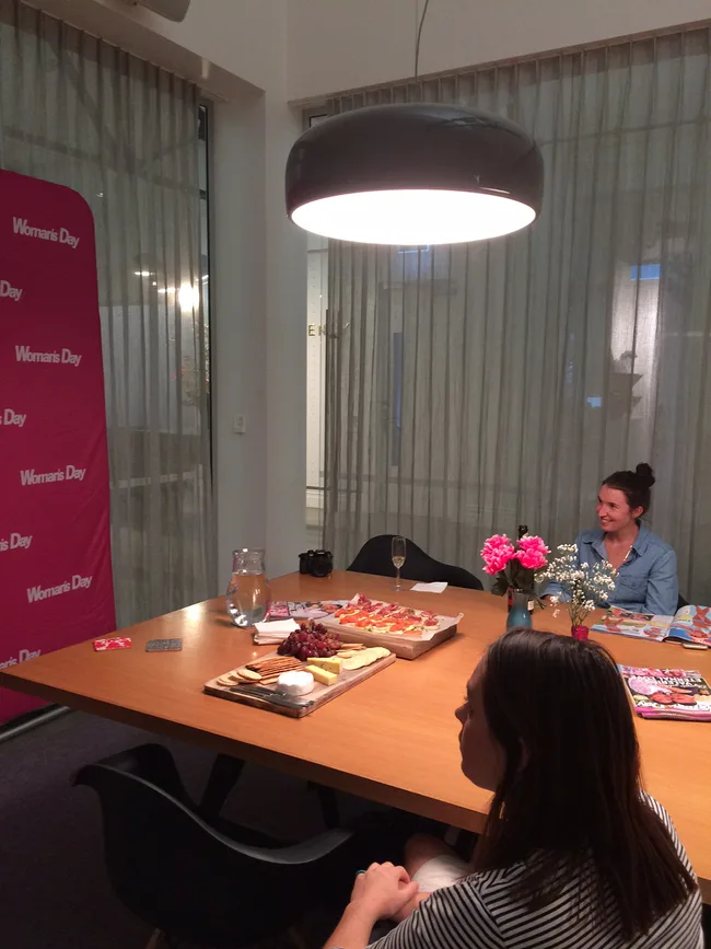 Two women sitting at a table with snacks and magazines, under a pendant light, with a "Woman's Day" backdrop.