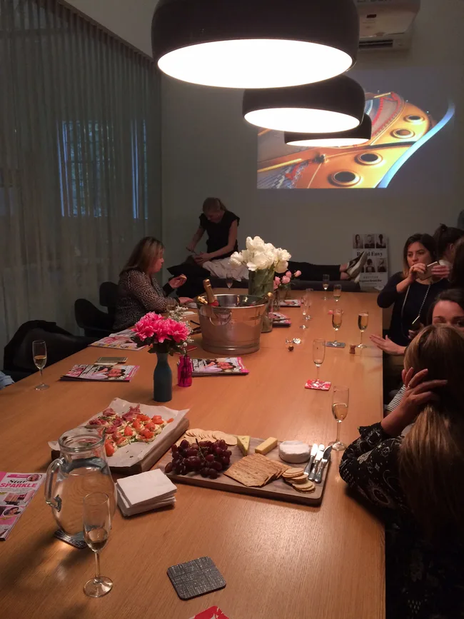 A group of people sit around a table with snacks, magazines, and glasses of champagne, under hanging lights.