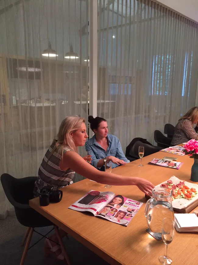 Two women sitting at a table, one reaching for food. Magazines and drinks are on the table in a modern setting.