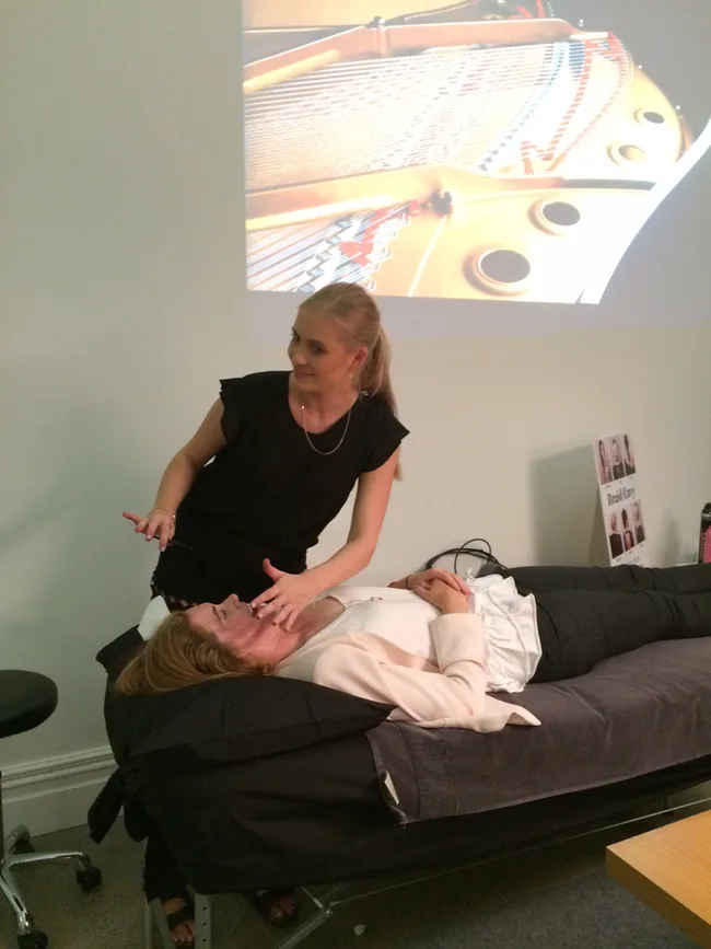 A woman receives a facial treatment, lying on a table, with a technician applying a product; piano image projected behind.