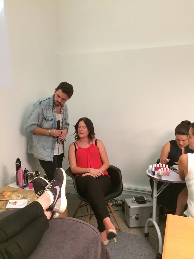 Three people are in a room; one is seated in red, one is standing, and another is doing nails at a small table.