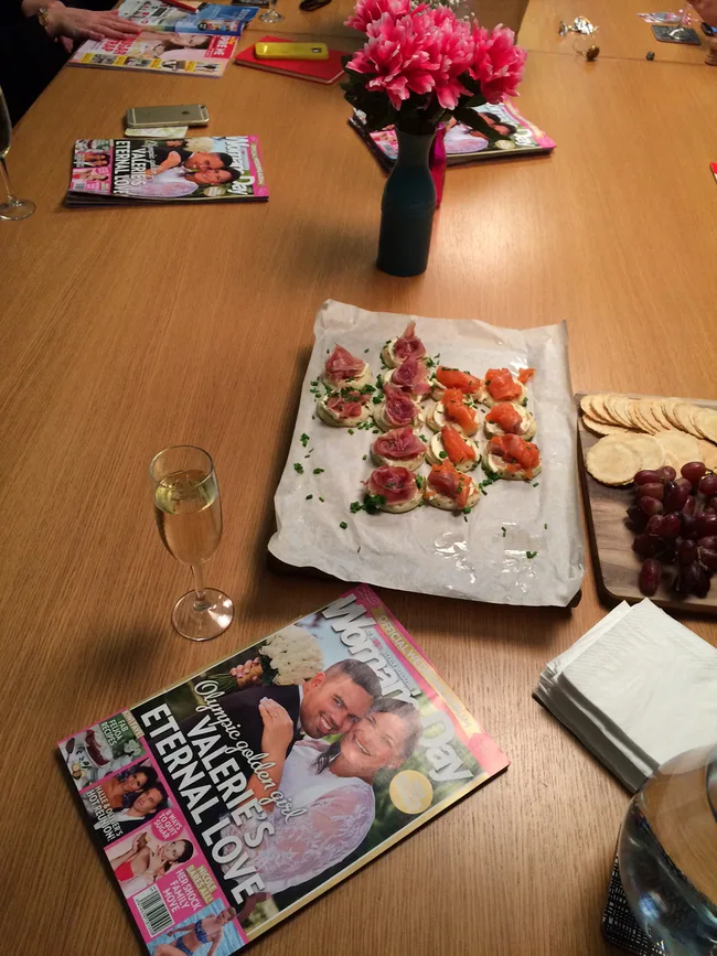 A table with a magazine, appetizers, a glass of champagne, and a vase of pink flowers.