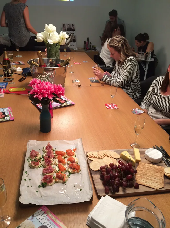 A group of people sitting around a table with flowers, party snacks, and champagne bottles in an ice bucket.