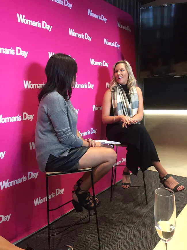 Two women sitting and talking in front of a pink "Woman's Day" backdrop.